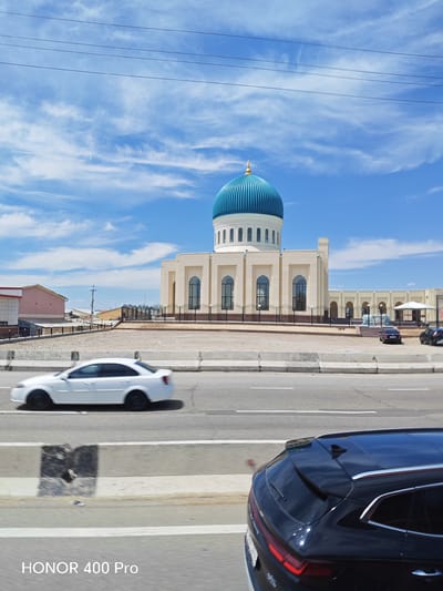 A mosque with a turquoise dome and beige facade is visible in the background under a partly cloudy blue sky, with power lines strung across the top of the frame. In the foreground, a white sedan is captured in motion on a highway, blurred due to its speed. To the right, the rear of a dark SUV is also visible on the highway. The setting appears to be a suburban area, possibly on the outskirts of Yangi Chinaz, Uzbekistan, indicated by the architectural style of the mosque and surrounding buildings.