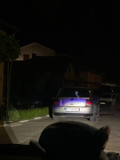 The image depicts a street scene at night in the city of Yasen, Bulgaria. In the foreground, a dark, out-of-focus object, possibly an animal's head or a piece of furniture, is visible. The middle ground is dominated by the rear of a silver Volkswagen Golf car, its license plate reading "EH 9488 KC". The car's taillights are illuminated, and a wispy white cloud, possibly exhaust or steam, is visible above its rear. To the right of the Golf, a lighter colored car is parked. The background shows the dark facades of buildings with dimly lit windows. A green hedge runs along the side of the road in front of the buildings. The street is paved and appears to be sparsely lit, with some scattered white debris on the surface. The overall atmosphere is nocturnal and somewhat muted due to the low light conditions.