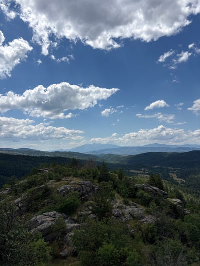 The image depicts a panoramic view of a mountainous landscape, likely in the vicinity of Dolno Drianovo, Bulgaria. The foreground is dominated by a rocky outcrop covered in lush green vegetation, including trees and bushes. Large, weathered boulders are scattered across the terrain, suggesting a natural, rugged environment.
Beyond the immediate foreground, the landscape unfolds into a series of rolling hills and valleys, densely covered with dark green coniferous forests. In the mid-ground, the forest gives way to more open areas with scattered fields and what appears to be a small village nestled among the trees. Distant mountain ranges are visible on the horizon, fading into a hazy blue, indicating a considerable distance.
The sky above is a vibrant blue, punctuated by a dramatic display of white, puffy cumulus clouds. Some of these clouds are large and dense, casting shadows on the landscape below, while others are smaller and more scattered. The sunlight suggests it is daytime, likely mid-morning or early afternoon, given the brightness and the angle of the light. There are no people or discernible man-made structures in the immediate foreground or mid-ground, beyond the hint of a village in the distance. The scene is one of natural beauty and tranquility.