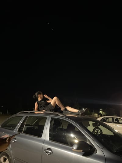 A person in a black sleeveless dress and black shorts is lying on the roof of a silver station wagon at night. The person's legs are extended, and they are looking to the left with their right hand near their head. The car is parked in what appears to be an outdoor, possibly gravel, area. In the background, another car, a white sedan, is visible, along with some sparse vegetation and lights from buildings. The sky is dark and clear, with a few distant lights visible. The overall impression is of a casual, possibly clandestine, gathering in a semi-urban or rural setting. The location is identified as Limenas Avdiron, Greece.