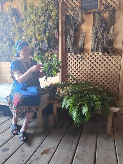 A woman wearing a blue headscarf and a blue patterned dress sits on a wooden bench, holding a bundle of green leafy branches. In front of her, on the bench and the wooden floor, are piles of dried herbs, ferns, and small white flowers. Behind her, the wall is adorned with more dried herbs hanging from the ceiling and wooden lattices. The floor is made of wooden planks. The scene appears to be an interior space, possibly a rustic outdoor building or a dedicated herb drying area, within the city of Mokhovichki, Russia.
