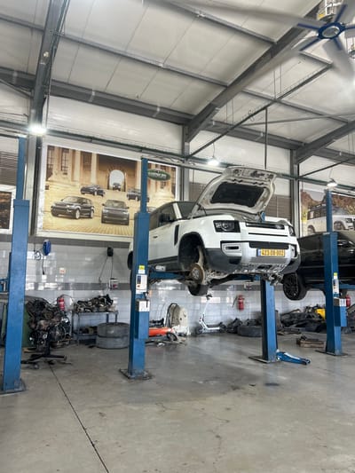 An auto repair shop features a white Land Rover Defender elevated on a blue lift. Its hood is open, revealing the engine bay. Car parts and tires are scattered around the shop floor. A poster showcasing various Land Rover models adorns the wall. Another car is being worked on nearby. The shop's interior includes a metal roof structure and bright lighting.