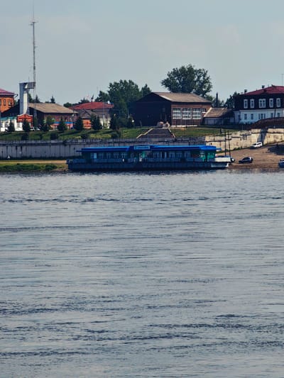 The image shows a wide, calm river in the foreground. In the middle ground, a green and blue boat or ferry is docked at a concrete embankment. Beyond the embankment, a city skyline is visible, featuring various buildings, trees, and a tall radio tower. The architecture suggests a historical or provincial Russian setting. On the riverbank, there are a few cars parked, and a pedestrian walkway is evident. The sky is partly cloudy. The scene is likely a view of the city of Yeniseysk, Russia, situated on the Yenisei River, a major waterway. The overall impression is one of a tranquil day in a riverside town.