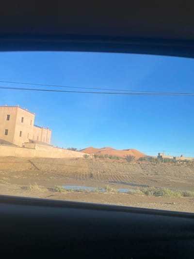 Captured from inside a vehicle, a desert landscape unfolds. A light brown building stands adjacent to a sandy hill. The terrain shows signs of tire tracks and small vegetation. A clear blue sky stretches above, intersected by power lines. The scene suggests a journey through an arid region.
