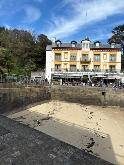 The image shows a view of the Hotel de la Vallee in Dinard, France (latitude: 48.636, longitude: -2.049). The hotel, a three-story yellow building with a red roof, is located on the waterfront, facing a sandy beach.  The beach is currently at low tide, exposing a significant portion of the sand.  A stone wall separates the hotel from the beach.  A group of people stand on the walkway in front of the hotel, including several people standing on the patio in front of the restaurant.  Several vehicles are parked in front of the hotel. The sky is clear with scattered clouds, the sun shines brightly, and the weather appears pleasant. The image was likely captured during the spring or summer months.