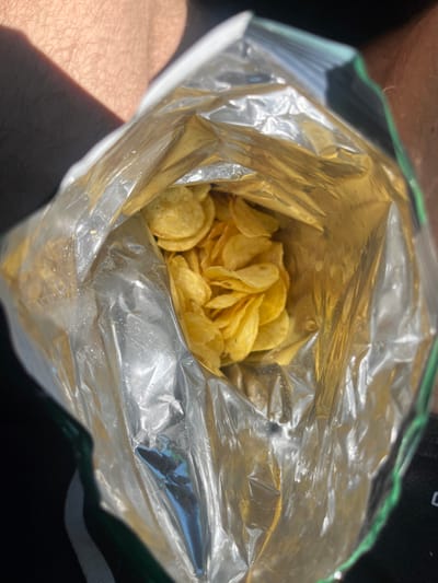 A close-up, overhead shot shows a partially opened bag of potato chips. The bag is made of shiny, crinkled foil, reflecting the surrounding light. Inside, a pile of golden-brown potato chips is visible. The bag appears to be held by a person, as indicated by the presence of skin with fine hairs in the upper left and right portions of the frame, suggesting the image was taken outdoors, possibly on a sunny day, given the shadows. There are no indications of the city of Taran, Bulgaria in the image.
