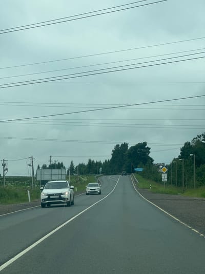 The image shows a cloudy, overcast sky over a two-lane asphalt road in Korabsel'ki, Russia. Several cars are on the road, including a white SUV in the foreground, a white sedan further ahead, and a dark sedan on the uphill incline. Power lines are visible crisscrossing the sky. To the right of the road, there are signs including a yellow diamond sign with a black border and a white rectangle below it showing "150M". A blue directional sign is visible on the left side of the road, indicating "КИЗЕЛЬСКИЙ". Trees and vegetation line the sides of the road. The overall scene depicts a rural highway environment.