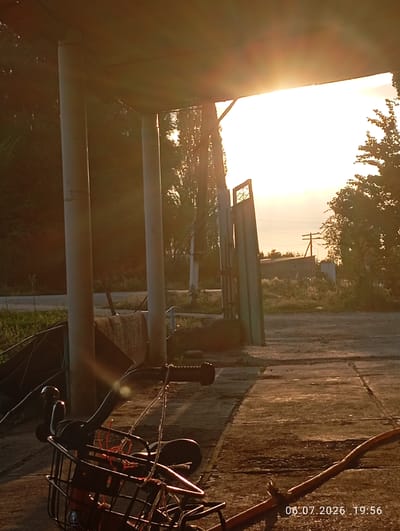 The image depicts an outdoor scene in a rural or semi-rural area, possibly within or near the city of Кызыл-Сай, Kyrgyzstan, as indicated by the timestamp. The perspective is low, looking upwards towards a bright, sunlit sky, suggesting it is either sunrise or sunset.
In the foreground, the handlebars and front basket of a bicycle are visible. The basket is empty, and some red twine is loosely tied within it. A wooden stick lies diagonally across the lower right portion of the frame. The ground appears to be paved with concrete slabs, some of which are cracked or uneven.
Behind the bicycle, there are several large, cylindrical pillars supporting a structure that extends across the top of the frame. The sunlight strongly illuminates the area, creating lens flare and casting long shadows. Beyond the pillars, a metal gate or fence structure is partially visible on the right side of the frame.
The middle ground shows a clearing with some vegetation and a dirt or gravel path. In the background, there are trees, a telephone pole with wires, and a small, indistinct building with a sloped roof. The overall impression is one of a quiet, possibly abandoned or infrequently used location at the end of the day. There are no people visible in the image. The timestamp "06.07.2026 19:56" is imprinted on the lower right corner of the image.