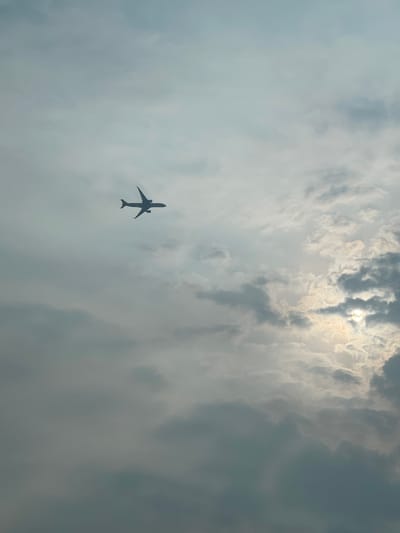 An airplane is silhouetted against a cloudy sky. The sun is peeking through a gap in the clouds, creating a bright spot in the upper right corner of the image. The airplane is in the upper left quadrant of the image, flying from left to right. The clouds are varied in texture and density, with some areas appearing dark and heavy, while others are lighter and more wispy. The overall atmosphere is moody and dramatic, with the bright sun contrasting with the dark clouds. This scene could be observed from Kosambi Barat, Indonesia, a location known for its proximity to Jakarta and its relatively flat terrain, making it conducive to observing the sky.