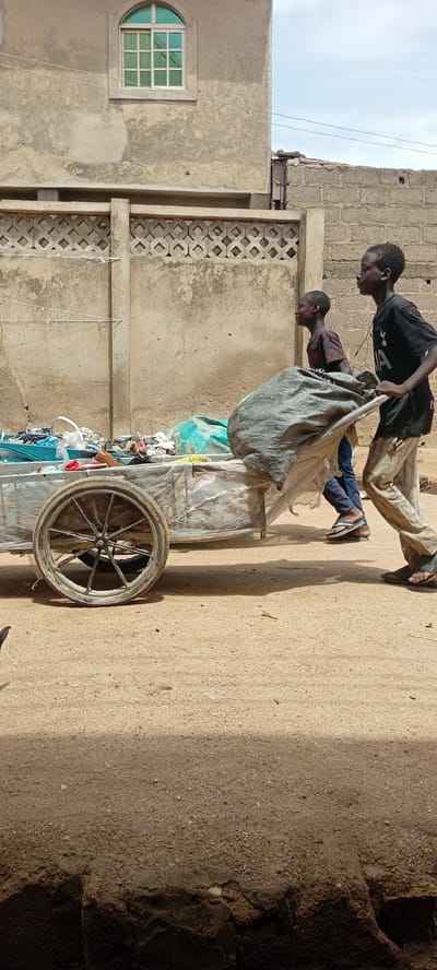 Two young boys are pushing a cart loaded with debris down a dusty dirt road in Rijiyar Zaki, Nigeria. The cart, a simple metal frame with a single large wheel, is covered in plastic sheeting and piled high with various discarded items, including what appears to be fabric and plastic bags. The older boy, wearing a black t-shirt and light-colored pants, is at the back, pushing. The younger boy, in a dark t-shirt and blue jeans, is in front, also contributing to the effort. Behind them, a textured concrete wall with a decorative pattern runs horizontally, and further back, a building with a distinctive arched window can be seen. The ground is dry and sandy, suggesting a hot, arid environment. The overall scene depicts a common sight in many urban areas, where individuals, often young, engage in scavenging or waste collection for a living.