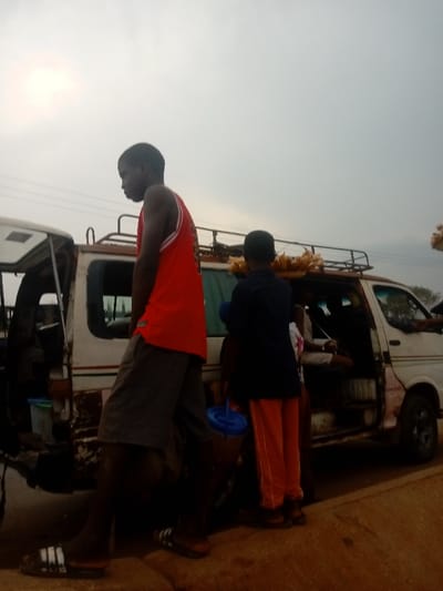Two people stand next to a white and brown van, likely a taxi or public transport vehicle. The person on the left wears a red basketball jersey, gray shorts, and Adidas sandals. The second person is wearing a dark jacket and orange pants. The van is full of people and has corn on the roof. The weather seems cloudy or overcast. The vehicle appears to be in poor condition.
