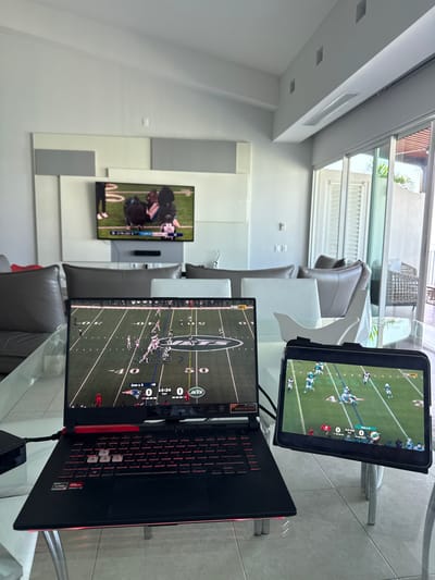 A modern living room features a large TV displaying a football game. On a glass table, a laptop and tablet also show football games, each displaying different teams and scores. The laptop shows the Jets and Patriots, while the tablet shows the Eagles and Dolphins. The setting is bright with natural light, illuminating the white walls and minimalist decor. Leather chairs surround the table, suggesting a comfortable viewing experience.
