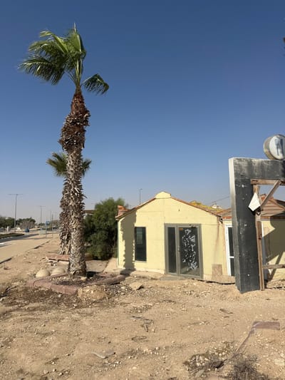 The scene is located in the Ramat Negev Regional Council, Israel, under a clear blue sky. The ground in the foreground and mid-ground consists of arid, sandy soil interspersed with small rocks and minimal dry vegetation. On the left, a tall palm tree with a textured trunk and green fronds stands within a low brick border. To its left, a paved pathway runs alongside a road with multiple streetlights and distant road signs visible. A single-story building with light-yellow stucco walls and a gabled roof is positioned in the central mid-ground. This building features a single vertical window on its left side and central double glass doors adorned with dark, abstract markings. Exposed roofing material is visible on the structure's roofline. To the right of the yellow building, a partially constructed or dismantled dark-colored structure with exposed wooden framing is present, displaying a circular sign with a fork and knife icon on its upper right side. Green foliage is visible behind the yellow building. No individuals are present within the frame, and no specific actions or interactions are depicted.