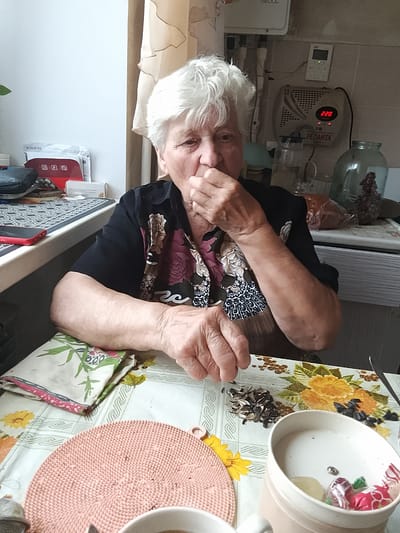 An elderly woman with white hair is sitting at a table in what appears to be a kitchen. She is wearing a dark shirt with a floral pattern. The table is covered with a tablecloth featuring a floral design. In front of the woman, there is a pile of sunflower seeds, some shelled and some unshelled, and a pink placemat. To her right, there is a white mug containing two candies and what appears to be a small coin. The background shows kitchen appliances, including a voltage stabilizer, and a large glass jar. The overall impression is one of a casual, home environment. The image was taken in Krasnoarmeysk, Russia.