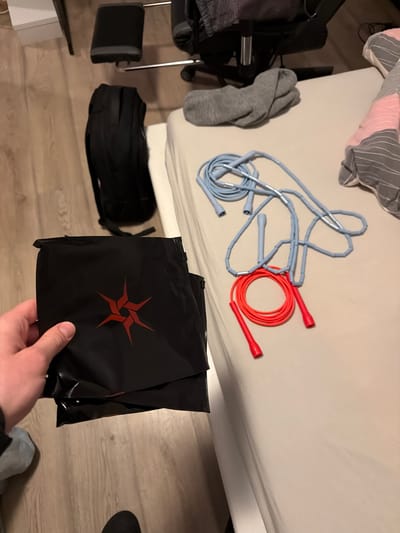 This high-angle indoor shot, taken in Benningen, Germany, captures a person's hand holding two black zippered pouches in the foreground. Each pouch features a distinct red, abstract, star-like logo, formed by four interconnected, inward-curving elements. On a light-colored bed, two jump ropes are displayed: a light blue beaded jump rope and a bright orange speed rope with red handles. Adjacent to the ropes, a grey sock and some pink and grey striped fabric are also resting on the bed. The scene includes parts of a black chair with a footrest and a black backpack on the light wooden floor to the left and bottom of the frame. The room is illuminated by what appears to be artificial light, typical of an indoor home setting. The image depicts a casual arrangement of fitness equipment and personal items within a domestic space.
