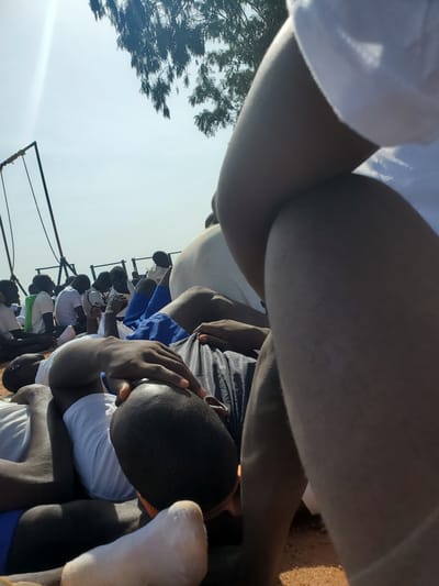In the city of Barakin Rafin Gora, Nigeria, a group of young men, likely students based on their uniforms of white shirts and blue shorts or trousers, are seen lying on the ground outdoors. The scene is characterized by a bright, possibly hazy, sky with sunlight filtering through the leaves of a tree overhead. A metal structure, resembling part of a swing set or some form of playground equipment, is visible in the background. The individuals are resting, some with their eyes closed, in what appears to be a relaxed or resting state. Their clothing suggests a communal setting, possibly a school or youth camp. The ground is covered in sand or dirt. The focus of the foreground is on the legs and arms of a person or people closest to the camera, with one individual's hand resting on their head.