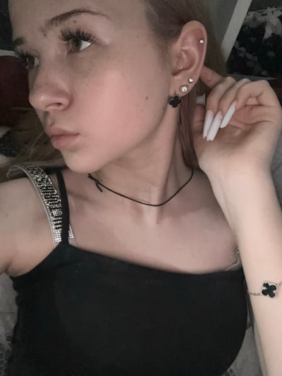The image shows a close-up, overhead shot of a young woman lying down. She has long, white acrylic nails and is wearing several pieces of jewelry: a black clover-shaped earring, a silver stud earring, and a silver bracelet with a black clover charm. She is also wearing a black top and a sparkly, rhinestone-embellished choker. Her face is turned away from the camera, with her eyes partially closed, giving a relaxed or contemplative expression. The lighting appears to be dim, suggesting it might be nighttime or indoors with low light. The background is blurred, but a hint of patterned fabric can be seen. The woman's hand is near her ear, with her fingers extended. The overall mood is intimate and personal.