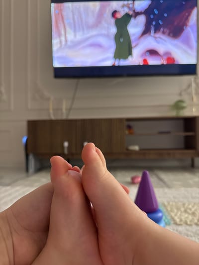 This image depicts an indoor domestic scene in Velikovechnoye, Russia, viewed from a low angle. In the immediate foreground, two light-toned, bare feet, consistent with those of an infant or young child, are positioned centrally and angled upwards. Below and to the right of the feet, a purple and blue conical plastic stacking toy rests on a light-colored, patterned rug. In the midground, a dark wood media console with a textured, possibly slatted, front and open shelving is visible. Above the console, a wall-mounted flat-screen television displays an animated scene featuring a figure in a dark green, long-sleeved garment with arms raised, set against a swirling backdrop of light purple, blue, and white hues dotted with small, bright elements. The wall to the left of the television features decorative paneling. A small, light green, spherical object is present on the right end of the media console. The environment appears to be a living area with ambient lighting.