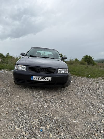 A dark-colored Audi A3 stands on a gravel path in what appears to be a rural or undeveloped area near Bornarevo, Bulgaria. The license plate number is PK 0655 KE. The sky is overcast. Vegetation is visible in the background. A small piece of blue trash is on the ground next to the car.