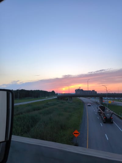The image shows a highway scene at sunset in Mirabel, Canada. A semi-truck with a dump trailer is traveling on the right lane of the highway. A white car is in the left lane ahead of the truck. A construction sign with an orange diamond shape and wavy lines indicating a bumpy road is visible on the right shoulder. In the background, a large building with multiple floors and a cell tower on top is situated beyond an overpass. The sky is a gradient of blue, transitioning to pink and orange hues from the sunset. A side-view mirror of a vehicle is partially visible in the lower left corner of the frame. Trees line the side of the highway.