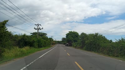 The image depicts a two-lane asphalt road in Lhokdagang, Indonesia, with a white line marking the left edge and intermittent yellow lines in the center. The road is flanked by dense green vegetation and trees on both sides. Utility poles with overhead electrical wires are visible along the left side of the road, with a cluster of them near the foreground. In the distance, a few vehicles are present on the road, including what appears to be a truck on the right side. The sky above is partly cloudy with patches of blue sky. The overall impression is that of a rural or semi-rural setting.
