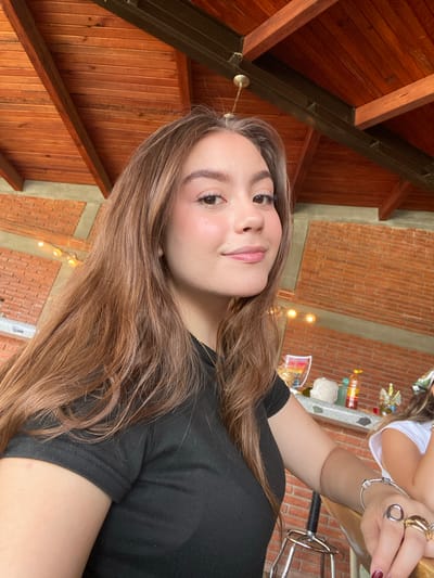 A young woman with light brown hair is indoors, in what appears to be San Francisco Lachigoló, Mexico, based on the architectural style. She is wearing a black t-shirt and looking towards the camera with a neutral expression. Behind her is a combination of brick and wood construction, suggesting a rustic or traditional building style. The lighting is warm, likely from natural light and possibly some artificial fixtures. To the upper right of the woman, partially out of focus, are a few hands adorned with rings and a bracelet. The setting appears casual, potentially a dining or social area.