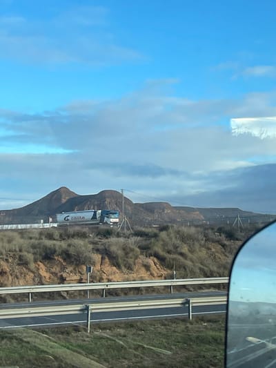 The image depicts an arid to semi-arid landscape viewed from a vehicle, with a portion of a side mirror visible on the right foreground. The sky is partially cloudy, featuring a mixture of bright blue and scattered white-to-light-gray stratocumulus clouds. In the immediate foreground, a multi-lane highway with a silver metal guardrail separates the viewer from the scene. Beyond the highway, an earthy embankment slopes upward, covered in sparse, dry, greenish-brown scrub vegetation and exposed reddish-brown soil. A small, dark rectangular signpost is embedded in this embankment.

Further into the midground, an elevated road or track runs horizontally, upon which a large white and blue articulated truck, branded with the "Costa" logo and a stylized 'C', is visible, traveling from left to right. The truck appears to be a tanker or bulk carrier. Adjacent to this road, a metal power pylon with overhead lines is present on the left, with additional pylons and lines extending across the middle distance.

The background is dominated by a series of rounded, barren hills or small mountains, exhibiting colors ranging from dark brown to reddish-brown, suggesting a dry, rocky geology typical of the region. This scene is situated in the vicinity of Fraga, Spain, consistent with the distinctive geological features and vegetation of the Monegros desert region. No individuals are visible.