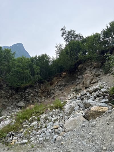 The image depicts a rocky, landslide-prone area with trees growing on the upper slopes. A mountain peak is visible in the background under a cloudy sky. The foreground consists of a steep incline covered in loose rocks and soil, with sparse vegetation. There are no people or animals visible in the scene. The location is Olden, Norway.
