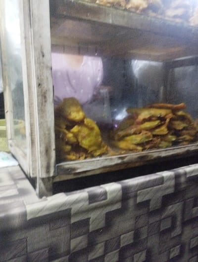 The image displays a close-up view of a food display case, likely situated at a food stall or small eatery in Kecamatan Jangkar, Indonesia. The central subject is a rectangular display case with glass panels and a worn, rustic frame, which appears to be made of wood or metal.
Inside the case, two distinct piles of deep-fried food items are visible. These items are yellowish-brown and appear to be fritters or pakoras; the pile on the left contains more irregularly shaped pieces, while the right pile consists of flatter, layered items. Above these, another layer of similar food items is faintly visible on a higher shelf within the display.
In the blurred background, partially seen through the glass and possibly as a reflection, is the torso of a person wearing a light-colored top, which appears to be pink or lavender with subtle white dot patterns. Their facial features are not discernible.
The display case rests on a counter or table with a distinctive patterned surface. This pattern features interlocking rectangular blocks in various shades of grey, white, and black, creating a tiled or geometric effect. The lighting in the scene is dim, suggesting it might be evening, night, or an interior setting lit by artificial light. There is no visible text in the image. The overall scene depicts a casual local food vendor setting.