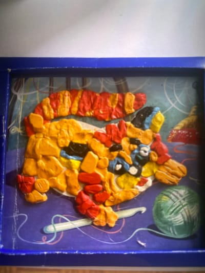 The image shows a colorful, textured artwork composed of what appears to be clay or play-doh. The artwork is framed by a dark blue border and is set against a purple background with abstract line drawings. The central subject of the artwork is a creature with a segmented, red and yellow body, resembling a caterpillar or a dragon. It has multiple limbs and a detailed head with black and blue elements that could represent eyes or facial features. To the lower left of the creature is a light blue crochet hook with a white handle, positioned as if it's part of the scene's narrative or simply an artistic element. To the lower right is a ball of green yarn with visible texture. The background features swirling lines in white, pink, and yellow, suggesting movement or a decorative pattern. The overall impression is whimsical and handmade.
The image does not provide any visual cues or context that would allow for the identification of its location in the city of Kerava, Finland.