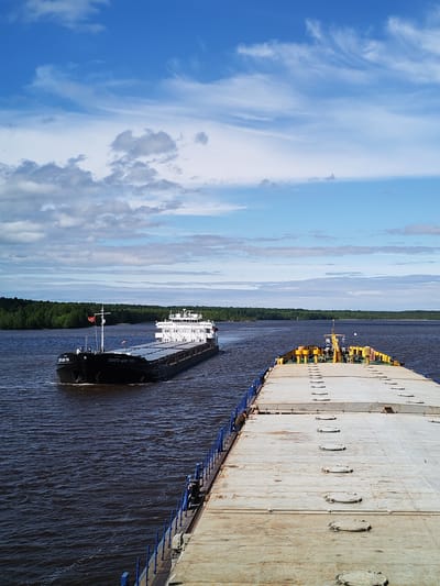 The image shows two barges on a wide river, likely the Volga. The barge in the foreground is light grey and flat, with a blue railing. The second barge is black, with a white superstructure and a red flag. The barge is labeled "ВОЛГО-ДОН 5551." The background shows a forested shoreline under a blue, partly cloudy sky. The location is likely near Popovo, Russia, given the Volga-Don designation on the barge.