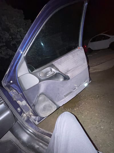 The image shows the interior of a dark-colored car door. The door is open, and the interior panel is a textured gray with a black door handle and speaker. A leg, clad in gray fabric, is visible to the right of the door. A vehicle with a red light, appears to be parked outside on the left. The surrounding environment appears to be a dark, unlit area with some vegetation visible outside the car window and a rough surface. The location is not identifiable as being in Kermen, Bulgaria, from the image alone.