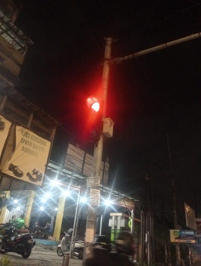 This media file depicts a street scene at night in Banjarkemantren, Indonesia. The main subject is a traffic light showing a red signal, casting a bright red glow on the surroundings. A utility pole supports the traffic light and a small electrical box.

In the foreground, several motorcycles are parked on the side of the road. To the left, a banner advertising "CICI GUDANG EPATU SAFETY BERMERK" (which translates to "Cici Warehouse Safety Shoes Branded") is displayed, featuring images of safety shoes. Further back, there are buildings with bright lights illuminating the area, suggesting a commercial or residential setting. People can be seen sitting at tables outside one of the establishments.

Visible text on signs includes "PANGGIL SERVICE" (Call Service) with a phone number, "TERIMA CATRIDGE BAR/LEKAS HARGA TINGGI" (Receive Cartridge New/Used High Price) with another phone number, and "AL PATTAN 15 MENIT" (Al Pattan 15 Minutes), possibly indicating a business name and service time. The presence of motorcycles, the type of buildings, and the Indonesian text on the signs strongly suggest the location is Indonesia. The dark sky and artificial lighting indicate it is nighttime. The overall atmosphere appears to be that of a lively street scene in an Indonesian town or city at night.