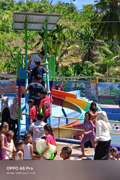 The image shows a group of children and adults at a water park in Palohseulimeng, Indonesia. A water slide with a ladder is in the center of the frame. Children are climbing the ladder to reach the top of the slide. Other children are gathered near the bottom of the slide, some holding a water gun. Adults are standing around, some wearing hijabs. In the background, a mural adorns a wall, and lush vegetation covers a hillside. The sky is blue and the lighting is bright, suggesting it is daytime.
