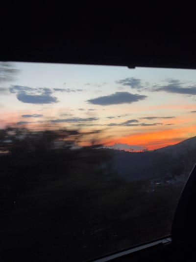 A photograph displays a sunset sky filled with clouds, transitioning from soft blues and grays to vibrant oranges. The sky meets a dark, indistinguishable mass on the right side, possibly representing land or trees. The picture is taken at an angle, suggesting it is an image of a screen or a framed print. The surrounding area is predominantly black, emphasizing the luminosity of the sunset.