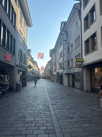 The image depicts a street scene in Winterthur, Switzerland, likely taken during the daytime under clear skies. The perspective is from a low angle, looking down a cobblestone street lined with multi-story buildings. Several flags, including the Swiss flag, are visible hanging across the street.
On the left side of the street, buildings feature numerous windows and architectural details. Signs for businesses are visible, including "metro" and "COZZIO." One sign advertises "50-80% AUF TOP BRANDS DAMEN HERREN KIDS IM 1.0G," indicating a sale on clothing for women, men, and children on the first floor of a store.
On the right side, the cobblestone pavement dominates the view, leading the eye down the street. A single person is walking away from the camera on the right side of the street. Further down the street, more buildings and flags are visible, suggesting a busy commercial area. The overall impression is of a pleasant, sunny day in a European city center.