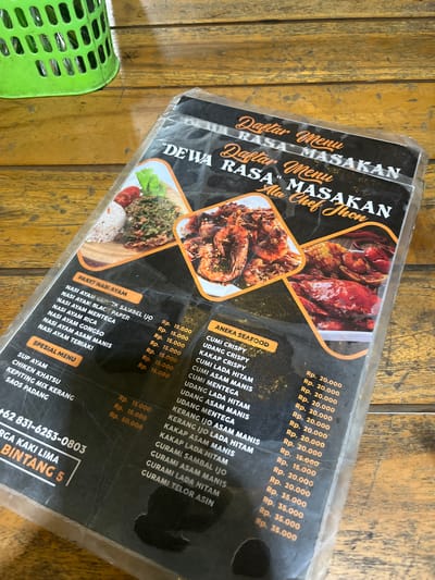 The image shows a laminated menu from "DEWA RASA MASAKAN Ala Chef Jhon" placed on a wooden table. The menu is partially folded and exhibits some glare from the lighting. To the left of the menu, a bright green, perforated plastic basket is visible, also resting on the table. The menu itself displays various food items categorized into "PAKET NASI AYAM", "SPESIAL MENU", and "ANEKA SEAFOOD". Under "PAKET NASI AYAM", items like "NASI AYAM CAPEK SAMBEL IJO" and "NASI AYAM BLACK PAPER" are listed with prices starting from Rp. 15.000. The "SPESIAL MENU" includes items such as "SUP AYAM" and "CHICKEN KHATSU". The "ANEKA SEAFOOD" section features dishes like "CUMI CRISPY" and "UDANG CRISPY", with prices around Rp. 20.000. Towards the bottom left of the menu, a phone number "+62 831-6253-0803" is displayed along with "IRGA KAKI LIMA BINTANG 5". The overall environment suggests a casual dining setting in Boja, Indonesia, as indicated by the menu's text and the likely context of an "IRGA KAKI LIMA" establishment.