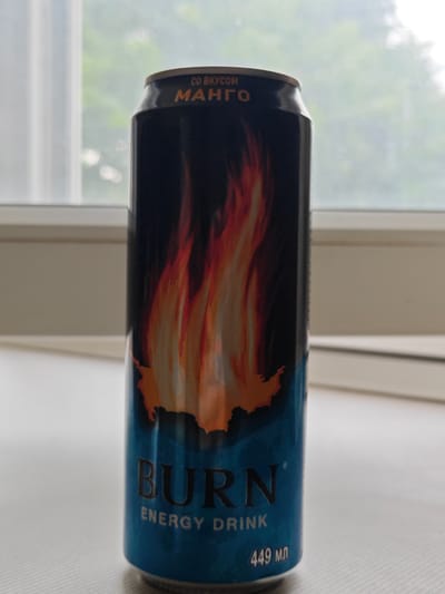 A black, blue, and orange "Burn" energy drink can with flames is positioned on a white surface in front of a window. The can has the text "СО ВКУСОМ МАНГО" at the top, and "BURN ENERGY DRINK" and "449 мл" at the bottom. The background outside the window appears to be foliage, suggesting an outdoor setting, possibly residential, given the window frame and sill. No people are present in the image, and no specific location within Dubovoye, Russia, can be identified from the visual information.