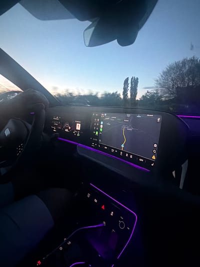 The scene captured is from inside a car, likely during twilight or early evening, with a focus on the dashboard and the view through the windshield. The dashboard features digital displays lit up, showing navigation, media information, and other car settings. The interior is accented with purple ambient lighting, giving it a modern feel. The person in the driver's seat is holding the steering wheel with both hands. Outside the car, the sky is visible, along with trees and possibly distant buildings or landscapes. The navigation system shows a route in the city of Mitino, Bulgaria.