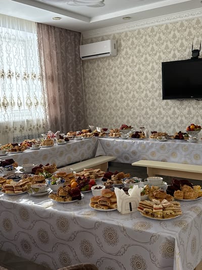 A long table laden with plates of assorted food, including sandwiches, cakes, and fruit, dominates the scene. The table is covered with a white tablecloth featuring a gold floral pattern. Napkins are stacked near the center of the table. A long, off-white cushioned bench runs along the side of the table. A window with sheer curtains and heavier patterned drapes is visible in the background. The ceiling has patterned wallpaper. A TV and an air conditioning unit are mounted on the wall. The location is Урмаево, Russia. No people are visible.