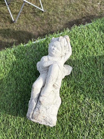 The image shows a stone statue of a person laying on its side, set on artificial grass. The statue is a light gray color, with visible weathering and texture. A tripod is visible in the lower left corner. The scene is located outdoors. The location in the city of Chirk, United Kingdom, is not discernible from the image.