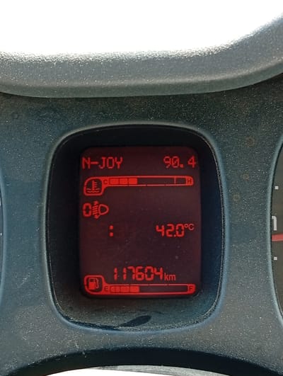 The image shows a close-up of a car's digital dashboard display. The display is illuminated in red and shows various indicators and readings. At the top left, "N-JOY" is displayed. To the right of "N-JOY" is the number "90.4". Below "N-JOY", there is a thermometer icon with waves, and a bar graph with several segments filled in, indicating temperature. To the right of the thermometer icon, "C" is visible, followed by a segment marked "H". Below the thermometer, there is an icon that appears to be a car headlight with a plus sign, and then a series of dots. Below this, the number "42.0°C" is displayed. At the bottom left, there is a fuel pump icon with a bar graph below it, indicating the fuel level, with "E" at the far left and "F" at the far right. To the right of the fuel indicator, the number "117604km" is displayed, indicating the mileage. The dashboard itself appears to be made of dark grey, textured plastic. The background of the image is blurred and suggests the interior of a vehicle. There are no people visible in the image. The image captures a moment from within a car, likely parked or stopped, showcasing the vehicle's operational status. The location in Chelopech, Bulgaria is not explicitly depicted or indicated in the image content.