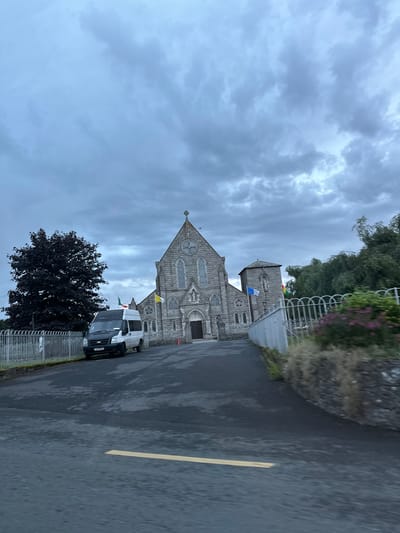 The image depicts a stone church with a prominent steeple, an entrance with large doors, and arched windows. In front of the church, a white van with a black front is parked on a paved area. Several flags are visible on poles near the church, including what appear to be the Irish flag and a yellow flag. To the left of the church, a dark-leaved tree stands beside a metal fence. The sky is overcast with heavy, dark clouds. The foreground shows a paved road with a yellow line marking. The overall scene suggests a religious building in an outdoor setting, possibly during inclement weather.