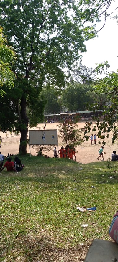 This image captures an outdoor scene in Tudor ward, Kenya, under bright daylight.
The foreground features a green grassy area covering the lower third of the frame, interspersed with drier patches and scattered light-colored litter. In the lower-left, two individuals are seated on the grass, viewed from behind; one wears a red shirt and the other a black shirt with white numbering "16". A light-colored concrete element is partially visible on the far right.
The midground is characterized by a large tree on the left with a thick trunk and dense green foliage, through which power lines are visible. Adjacent to this, a light brown, unpaved dirt field spans the central portion of the image. A rectangular, light-colored sign or board, appearing worn or damaged, stands on two posts near the field's edge, slightly left of center.
On the dirt field, a group of approximately 6-8 individuals, attired in matching orange athletic uniforms, are gathered near the sign, facing inwards towards the field. One adult in a light shirt and dark pants stands adjacent to this group. Further left on the field, several figures, some in yellow shirts, are engaged in a game. To the right, a person in a light blue-green shirt and dark shorts is observed running rightward while holding a light-colored, round object. Another small group of 3-4 individuals, dressed in white shirts and red shorts, stands further right on the field. An additional person is seated on the far right edge of the field.
The background displays a row of structures with dark green roofs, consistent with covered seating or shelters, positioned behind the dirt field. Beyond these structures and on the horizon, dense green trees are visible. The sky is bright white, partially obscured by the tree canopy.