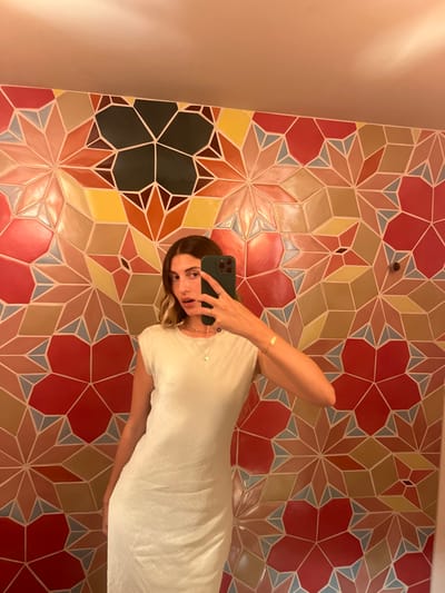 A mirror selfie captures a young adult female from the chest to mid-thigh, taken indoors at the Fondation Vincent van Gogh Arles in Arles, France.

The subject has medium-length, wavy brown hair with lighter ends. She is wearing a light-colored, possibly off-white, sleeveless midi-dress with a round neckline. A delicate gold-colored necklace with a small circular pendant is visible, along with a thin gold-colored bracelet on her left wrist. She holds a dark green smartphone, likely an iPhone, in her right hand, extended to capture her reflection in the mirror. Her mouth is slightly open, and her gaze is directed towards the camera lens.

The background features a distinctive, brightly colored tiled wall composed of repeating geometric patterns, including star-like shapes and polygons. The tiles incorporate a diverse palette of red, orange, yellow, pink, light blue, dark green, and brown hues, outlined by light-colored grout lines. A plain, lighter-toned upper wall or ceiling area is visible above the tiled section.