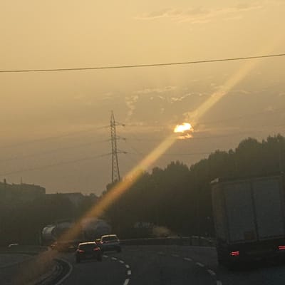 The image captures a view from a highway in El Vendrell, Spain, during what appears to be sunset or sunrise. The sky is a warm, hazy yellow, with scattered clouds. A prominent power line stretches horizontally across the upper portion of the frame. In the middle ground, a tall electricity pylon stands to the left. To the right of the pylon, the sun is partially obscured by clouds, casting a strong, diagonal beam of light that stretches downwards across the scene. Silhouetted against the hazy sky and foliage are trees forming a dark line along the horizon. On the highway, several vehicles are visible, including cars and a large truck. The road curves, and road markings are present. The overall impression is of a tranquil, albeit slightly hazy, moment on a highway as daylight fades or begins.