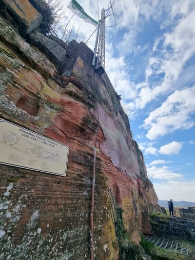 The image captures a sandstone rock formation, likely part of the Schlossberg Caves in Homburg, Germany. The rock face is weathered and covered in patches of moss and lichen. A metal tower structure extends upwards from the left side of the rock. A flag with green and white is attached to the tower. An information panel describing the site's history, specifically referencing the "Ehemalige Festung Homburg 1679-1697 * 1705-1714" (Former Fortress Homburg), is mounted on the rock face. A metal railing runs horizontally across the top of the rock formation. In the upper right corner of the image, a person is visible standing on the rock. The sky is blue with scattered clouds.