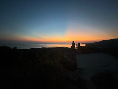 This media file captures a serene twilight scene in Karimunjawa, Indonesia. The image is dominated by a vast sky transitioning from a deep, dusky blue at the zenith to vibrant hues of orange and yellow near the horizon, indicating sunset or sunrise. Distinct sun rays fan out from a point low on the horizon, creating a dramatic and peaceful atmosphere.
In the foreground, silhouetted against the colorful sky, are two stone statues. The larger statue appears to be a depiction of a figure, possibly a Buddha, with a smaller, tiered structure beside it, resembling a pagoda or stupa. These statues are placed on a rocky outcrop overlooking a body of water, likely the sea, which stretches towards the horizon. Scattered lights are visible on the water, suggesting boats or distant settlements.
To the right, the silhouette of a hilly coastline is visible, adding depth to the landscape. The foreground is dark and shadowed, with the outlines of foliage and rocks suggesting a natural, elevated vantage point. The overall mood is tranquil and contemplative, amplified by the fading light and the quiet presence of the statues. The time of day is clearly twilight, with the last vestiges of daylight painting the sky. No text is visible in the image.