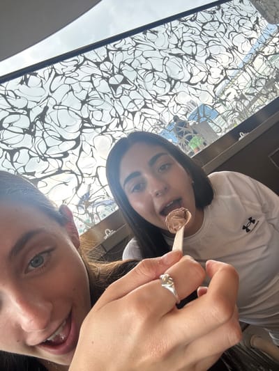The image captures two young adult females indoors at a location in the Berry Islands, Bahamas. In the foreground, partially visible on the left, is a female with blue eyes and a smile. Her right hand, featuring a silver knot-design ring, holds a wooden spoon with a scoop of chocolate-colored ice cream. This spoon is extended towards a second female, positioned centrally, who has dark hair and dark eyes. She wears a white long-sleeved shirt with a black Under Armour logo on the left chest and is shown with her mouth open, leaning forward to consume the ice cream. Overhead, a distinctive glass ceiling with an intricate, dark, organic-patterned metal overlay is visible. This ceiling reflects a bright sky with clouds and a blurred figure of an individual in the distance, suggesting an open-air or atrium-like environment under daylight conditions.