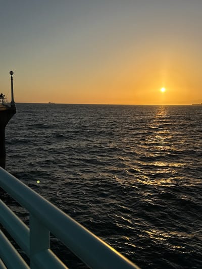 The image depicts a sunset over the Pacific Ocean, viewed from the Manhattan Beach Pier in Manhattan Beach, California. The sun, a bright orb, is positioned in the center-right of the frame, casting a golden-orange glow across the sky and shimmering path on the dark, rippling water. On the left side of the image, a silhouetted figure stands on the pier, holding a fishing rod. A lamppost with a circular light fixture is also visible on the pier. In the distance, on the horizon, several cargo ships can be seen. The foreground features the white railing of the pier.